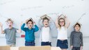Cheerful girls and boys stand in front of a chalkboard, each holding an open book over their head.