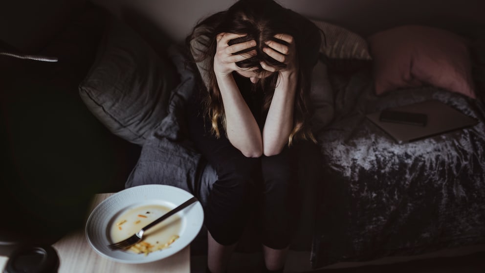 A woman sits on the bed holding her head. Next to her is an empty plate.