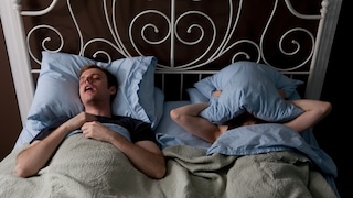 Pill for Sleep Apnea? A man lies snoring next to a woman in bed, who has placed a pillow over her head.