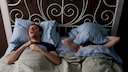 Pill for Sleep Apnea? A man lies snoring next to a woman in bed, who has placed a pillow over her head.
