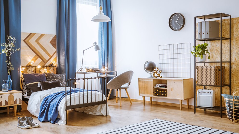 A teenager's room with a bed, desk by the window, a carpet and open shelves