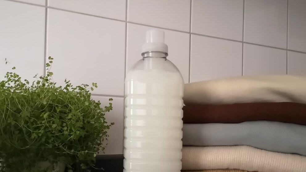 Laundry detergent stands in front of a pile of clothes and a plant.