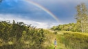 A child and a dog run across a green meadow as a rainbow appears in the sky.