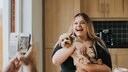 A woman cradles her poodle like a baby in her arms. Someone captures the moment on their phone.