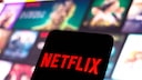 Netflix logo on a phone, with streaming tiles in the background.