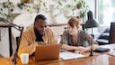 Two people are sitting at the table in front of a laptop.
