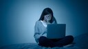 A woman sits on the bed with a bandaged hand and a laptop on her lap.
