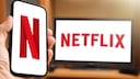 A person holds a phone displaying the Netflix logo, with a TV in the background showing the streamer's name.