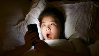 A girl lies in bed, looking startled at her smartphone.