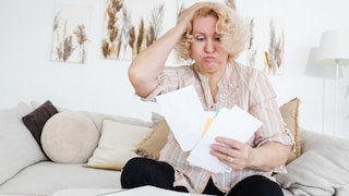 An elderly woman sits on a couch, looking stressed as she stares at a handful of letters in front of her.