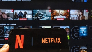 Smartphone with a large Netflix logo on the display. In the blurred background, various movie and series covers are visible.