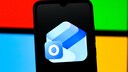 Smartphone displaying the Outlook app logo on a black screen, set against a background of red, green, blue, and yellow color blocks in the style of the Microsoft logo.