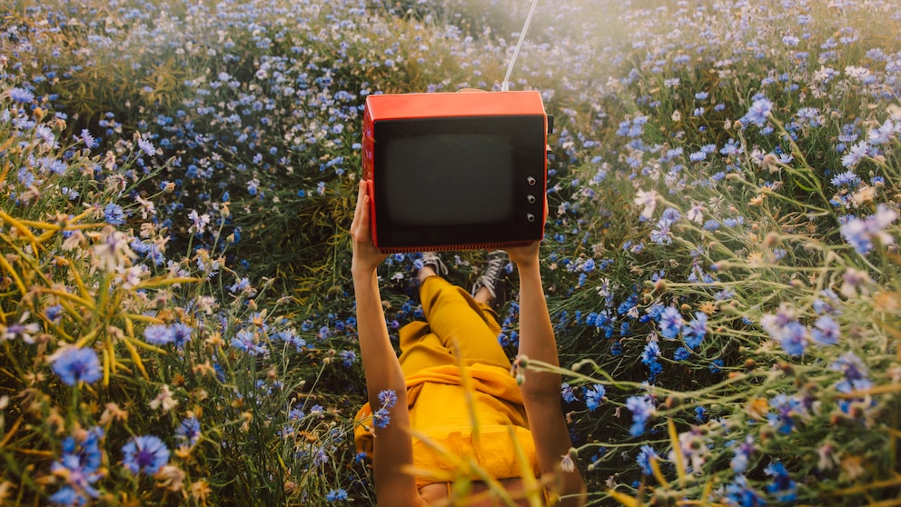 A person is lying in a meadow filled with numerous blue flowers, holding a red television with an antenna in front of their face. The person is dressed in yellow clothing and is lying on their back. In the background, there is a field, trees, and several wind turbines. The image was taken in warm daylight.
