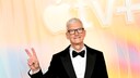 Apple's CEO Tim Cook stands against a yellow-pink background, flashing the peace sign.