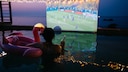 Person in Pool Watches Soccer on Big Screen