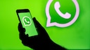 A hand holds a smartphone with a green screen displaying the WhatsApp logo. The background is the same green, featuring another WhatsApp logo.