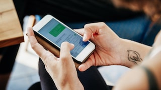 A person is holding a smartphone with both hands, typing on the on-screen keyboard. The display shows an open chat application with a green message bubble. The smartphone is in a protective case. A small tattoo is visible on the person's right wrist. In the background, other people and a wooden table can be seen, though they are out of focus.