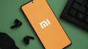 A smartphone with the Xiaomi logo on a green background. Next to it, the corner of a laptop and loose in-ear headphones.