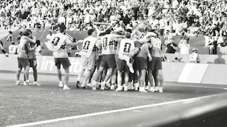 A black-and-white photograph captures a jubilant soccer team on the sidelines of a stadium. Several players and substitutes have gathered in a huddle, celebrating in front of the advertising boards along the field's edge. In the background, the stands are packed with numerous spectators.