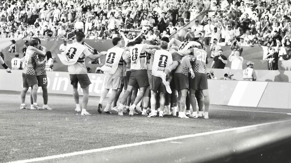 A black-and-white photograph captures a jubilant soccer team on the sidelines of a stadium. Several players and substitutes have gathered in a huddle, celebrating in front of the advertising boards along the field's edge. In the background, the stands are packed with numerous spectators.