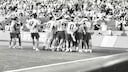 A black-and-white photograph captures a jubilant soccer team on the sidelines of a stadium. Several players and substitutes have gathered in a huddle, celebrating in front of the advertising boards along the field's edge. In the background, the stands are packed with numerous spectators.