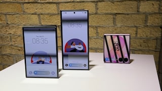Three Samsung foldable smartphones are displayed on a white table against a light stone wall. On the left is an unfolded device in landscape orientation with a large display. In the center, there's an unfolded device in portrait orientation with narrower screen borders. On the right, a partially folded pink smartphone is visible. All screens show the time as 8:35. The two larger devices also display an illustration on the lock screen. The photo was taken indoors with warm lighting.