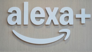 White 3D letters spelling "alexa+" are mounted on a light wooden surface. Below the lettering is the curved Alexa symbol in white. The image exclusively features the logo against a light wooden background.