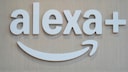 White 3D letters spelling "alexa+" are mounted on a light wooden surface. Below the lettering is the curved Alexa symbol in white. The image exclusively features the logo against a light wooden background.