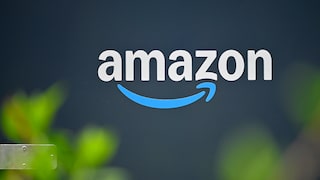 Amazon logo in white lettering on a dark blue background. Below the text is the company's blue arrow. In the foreground, blurred green plants are visible.