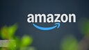Amazon logo in white lettering on a dark blue background. Below the text is the company's blue arrow. In the foreground, blurred green plants are visible.