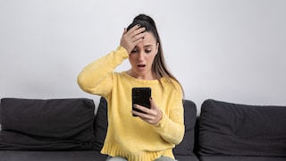 A person in a yellow sweater sits on a dark sofa, holding a smartphone in one hand. The other hand rests on their forehead, with a bright wall visible in the background.