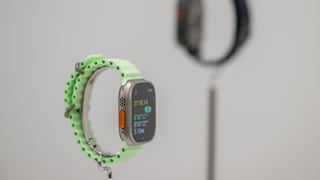 Apple Watch Ultra 3 with a light green band on a stand, with another smartwatch blurred in the background.