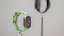Apple Watch Ultra 3 with a light green band on a stand, with another smartwatch blurred in the background.
