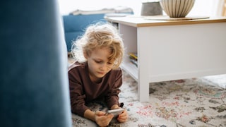 A child lies on a carpet and looks at a smartphone in a living room.