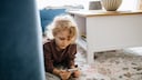 A child lies on a carpet and looks at a smartphone in a living room.