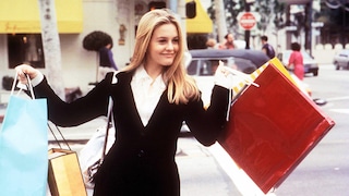 A person stands on a street holding several large shopping bags in various colors in both hands. In the background, blurred cars, shops, and other people can be seen.
