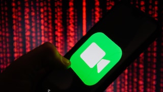 A hand holds a smartphone displaying a large FaceTime icon on the screen. In the background, blurred red light patterns evoke the appearance of digital data streams.