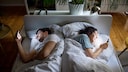 Two people lie back-to-back in a double bed, each looking at a smartphone. The bed is in a bright bedroom with hardwood floors, houseplants, and a bedside lamp. The light from the smartphone screens illuminates their faces.