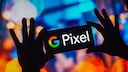 Two hands hold a smartphone in landscape mode against a blurred, brightly lit background. The display shows the colorful Google logo and the word "Pixel." Blue, orange, and violet light areas and bokeh effects dominate the background. The hands and smartphone appear as dark silhouettes against the bright colors.