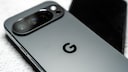 Close-up of a gray Google Pixel smartphone showing the camera module and Google logo on the back, with another blurred smartphone in the background.