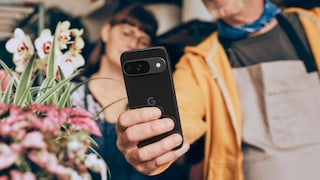 A person holds a black Google Pixel smartphone up to the camera. The device is sharply in focus in the foreground, with the Google logo and horizontal camera module clearly visible on the back. In the background, two blurred figures and several flowers and plants can be seen. The photo appears to have been taken indoors with warm lighting.