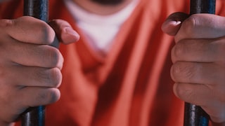 Close-up of two hands gripping prison bars. In the background, a person in orange clothing is blurred. The image symbolizes a prison situation.