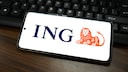 A smartphone lies horizontally on a dark wooden table in front of a black keyboard. The bright screen displays the blue ING logo and the bank's orange lion symbol. The smartphone is slightly angled and occupies most of the image.