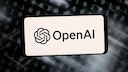 A smartphone or display shows the OpenAI logo and the word "OpenAI" against a bright background. In the background, there are blurred rows of zeros and ones, along with bright light streaks on a dark surface. The image resembles a graphic illustration related to artificial intelligence, software, and cybersecurity.