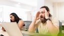 A person sits in a bright office in front of a laptop, holding their head in their hands. In the background, another person is seated at a laptop.