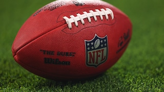 The image shows an American football with a clearly visible NFL logo on a grass field. The ball is in the foreground, while the background is blurred. This theme aligns with an article about the NFL's new TV rights.