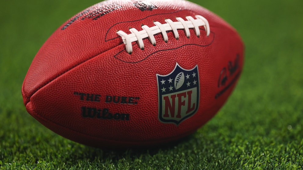 The image shows an American football with a clearly visible NFL logo on a grass field. The ball is in the foreground, while the background is blurred. This theme aligns with an article about the NFL's new TV rights.