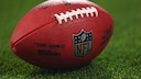 The image shows an American football with a clearly visible NFL logo on a grass field. The ball is in the foreground, while the background is blurred. This theme aligns with an article about the NFL's new TV rights.