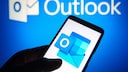 A smartphone is held against a blue-lit background featuring the Outlook logo. The display shows the Outlook app icon, which includes a blue envelope and a white "O." In the background, the word "Outlook" is blurred.