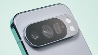 Close-up of the back of a silver smartphone featuring a large, oval camera module. Visible are multiple camera lenses, a flash, and additional sensors on a raised camera bar. The device is photographed at an angle against a bright, blurred background.